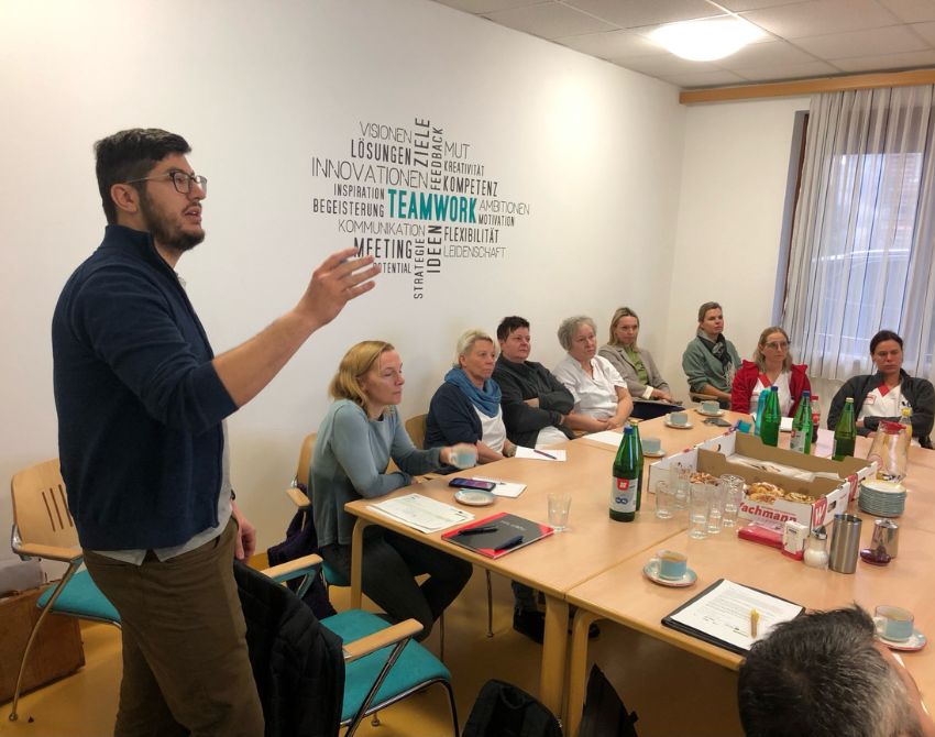 Teilnehmer:innen eines ready4heat Workshops sitzen um einen Tisch, im Hintergrund an der Wand das Wort "Teamwork". Dominik Puchner im Vordergrund steht und erklärt etwas.