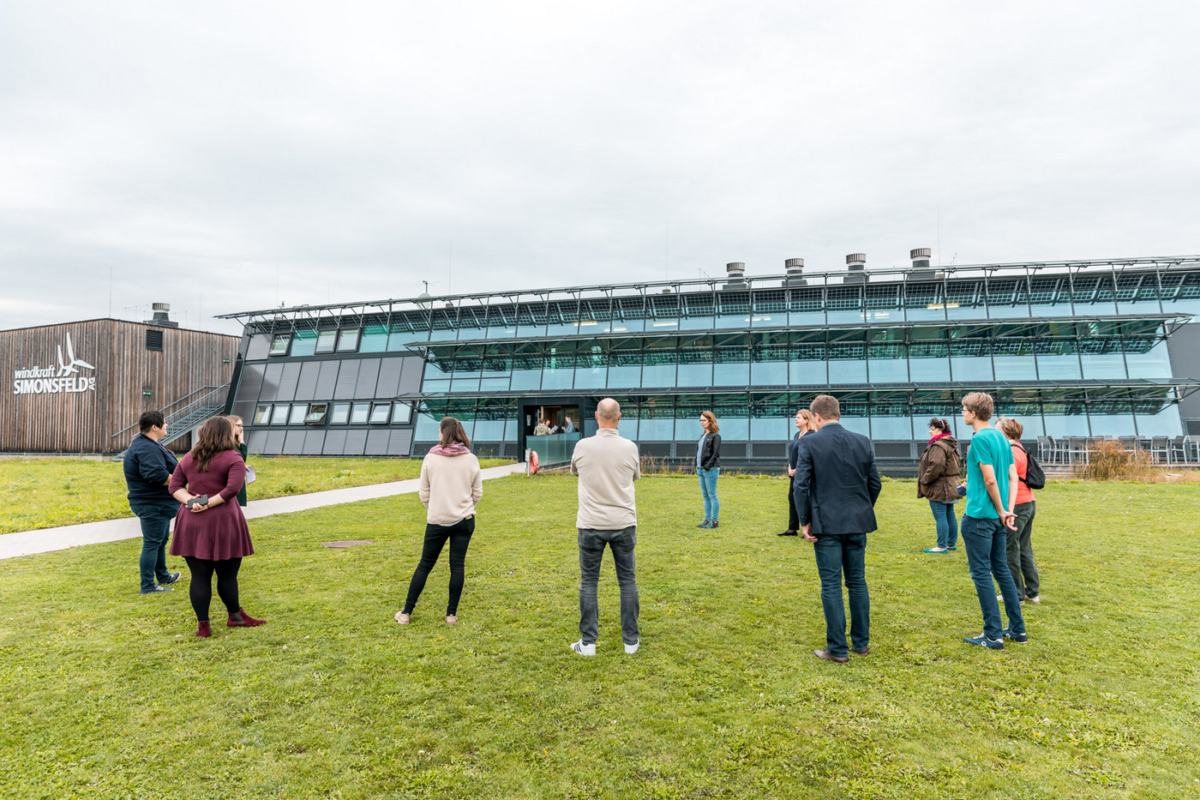 Eine Gruppe von Menschen stehen auf einer grüne Wiese vor einem Betriebsgebäude der Windkraft Simonsfeld.