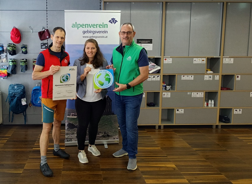 Zwei Männer und eine Frau stehen in einem Raum mit Kletterausrüstung und halten die Klimabündnis-Tafeln in der Hand.