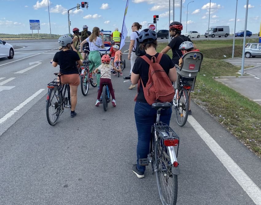 Eine Gruppe Radfahrende halten an einer Kreuzung an einer Landstraße, an der Autos fahren.