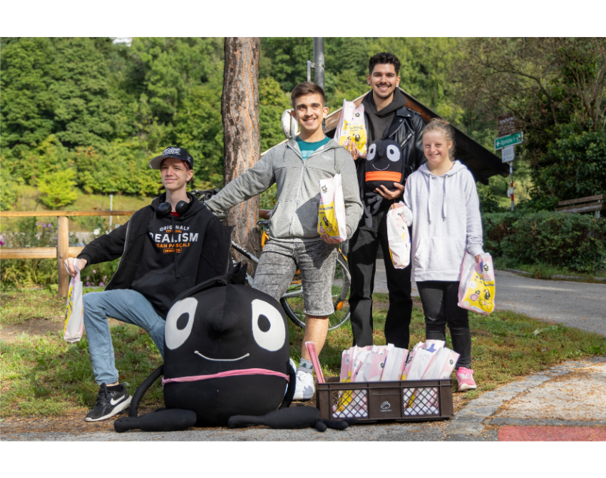 Vier Jugendliche stehen beisammen für ein Gruppenfoto mit dem Maskottchen Edgar während der Verteilaktion der Frühstückssackerl am Radweg in Innsbruck.