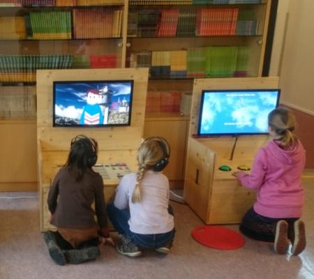 drei Kinder sitzen vor Stationen der KLIMAVERSUM Wanderausstellung vor Bildschirmen, mit dem Rücken zum:zur Betrachter:in