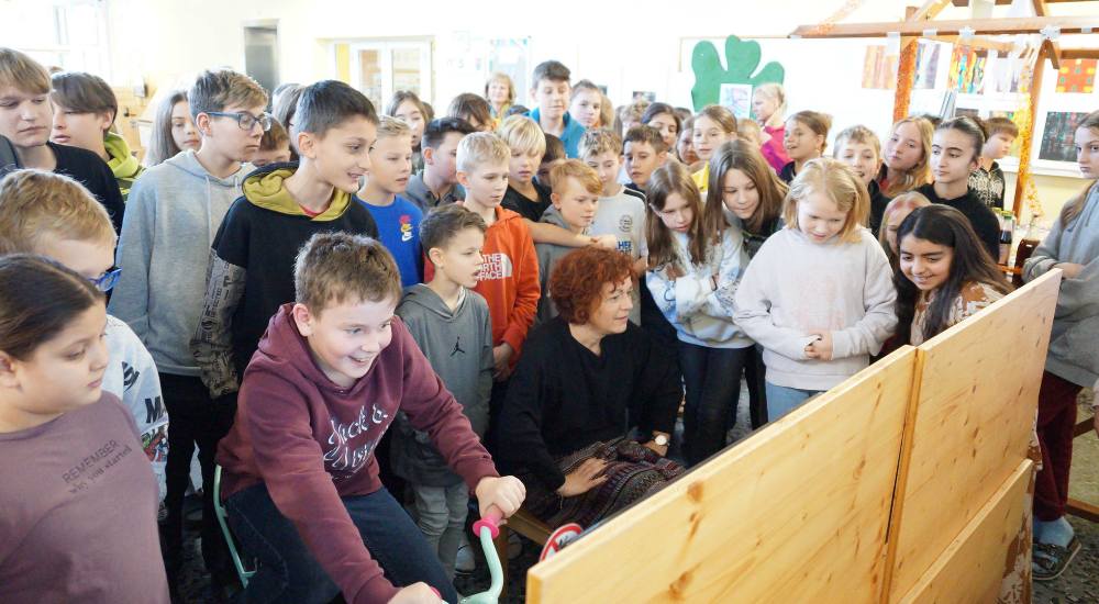 Cornelia Schweiner sitzt an der Station Verkehrsmittelvergleichsfahrt und Schüler:innen stehen um sie herum