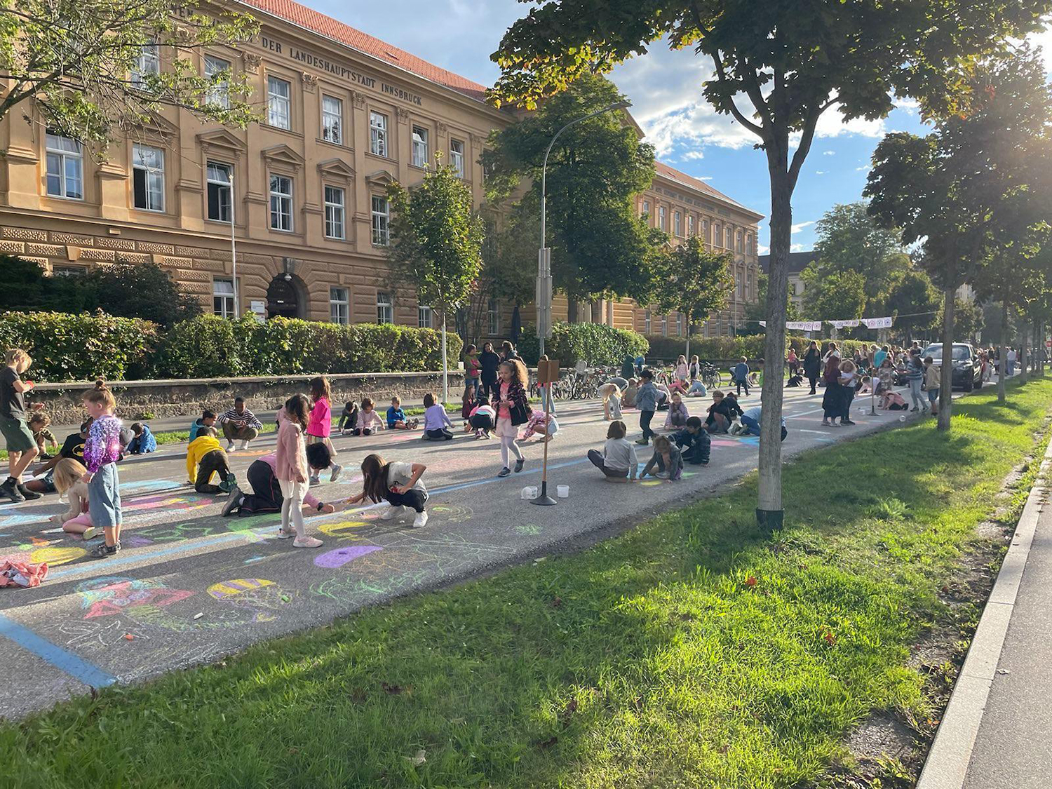 Eine Straße umsäumt von Grad und Bäumen, auf der Kinder mit Kreide bunte Zeichnungen malen.