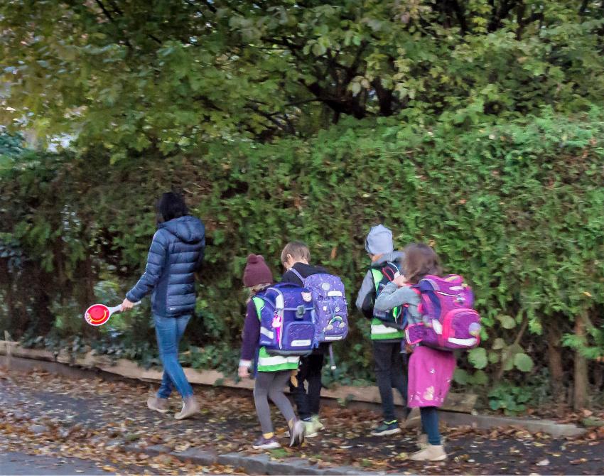 Eine Gruppe von Kindern geht hinter einer Erwachsenen auf dem Gehweg, dahinter eine Hecke