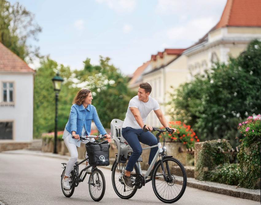 Eine Frau und ein Mann radeln der Betrachterin entgegen, schauen sich gegenseitig an, hinter ihnen Häuser und eine Hecke