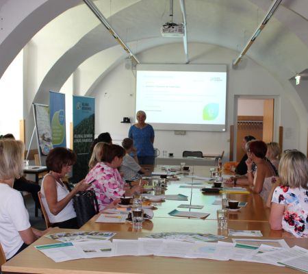 Teilnehmerinnen sitzen am Tisch und schauen auf eine Leinwand, vor der Steffi Greiter vom Klimabündnis Steiermark steht und referiert.