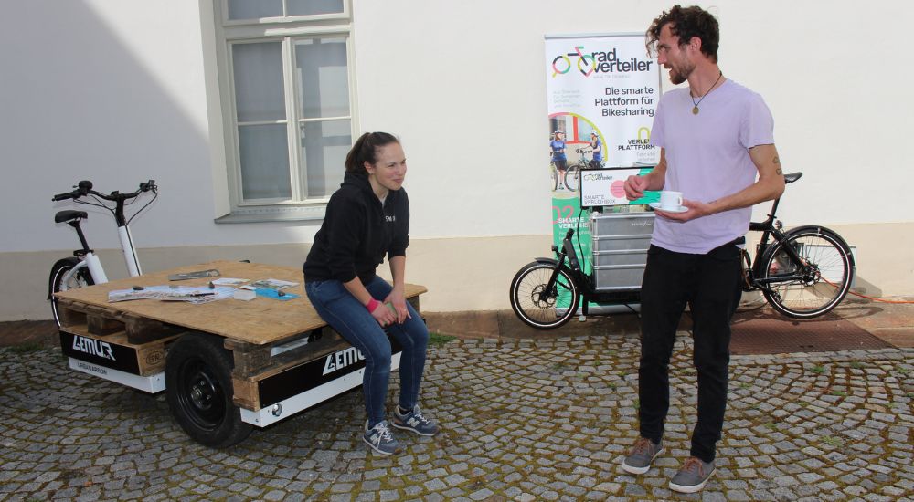 Frau sitz auf Ladefläche eines Lastenrads und unterhält sich mit neben ihr stehendem Mann.