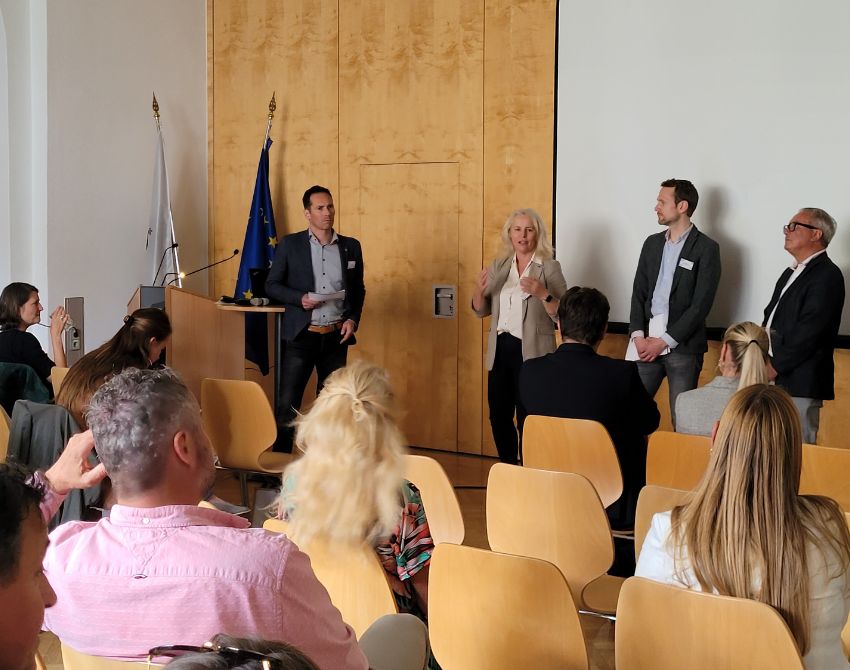 3 Personen stehen vor Leinwand und schauten auf vierte Person links von ihnen. Publikum schaut auf alle vier Personen.
