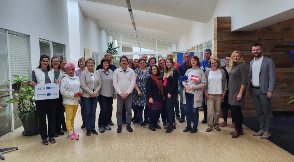 Gruppe von Erwachsenen posiert in hellem Innenraum, einige halten EU- und Österreich-Flaggen sowie Interreg-Schilder.“