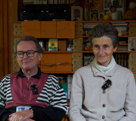 Ein Mann und eine Frau, beide älteren Alters, sitzen in ihrem Wohnzimmer und lachen in die Kamera