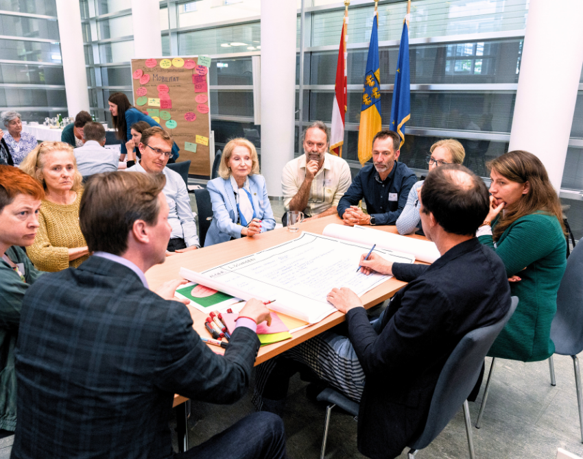 Experten sitzen rund um einen Tisch mit Flip-Chart und sprechen miteinander