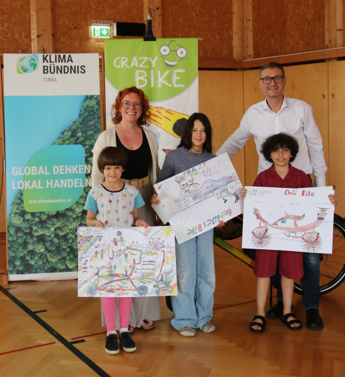 Drei Kinder halten Zeichnungen mit Fahrrädern. Hinter ihnen stehen zwei Erwachsene