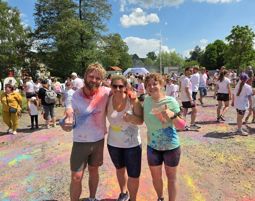 Zwei Frauen und ein Mann strahlen in die Kamera. Ihre Kleidung ist voller bunter Farbe.