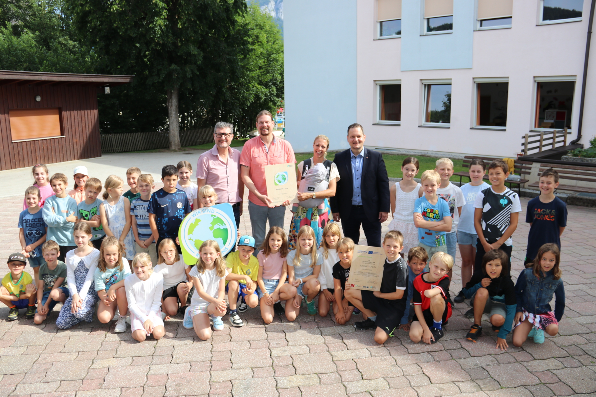 Kinder und Erwachsene sitzen und stehen auf Hof vor einem Haus. Einige halten Urkunden in der Hand.