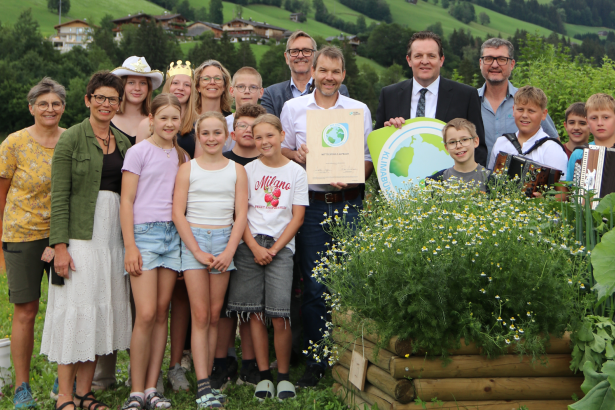 Fröhliche Kinder und Erwachsene im Grünen vor Wiesen und Bäumen mit Urkunden und Logo des Klimabündnis